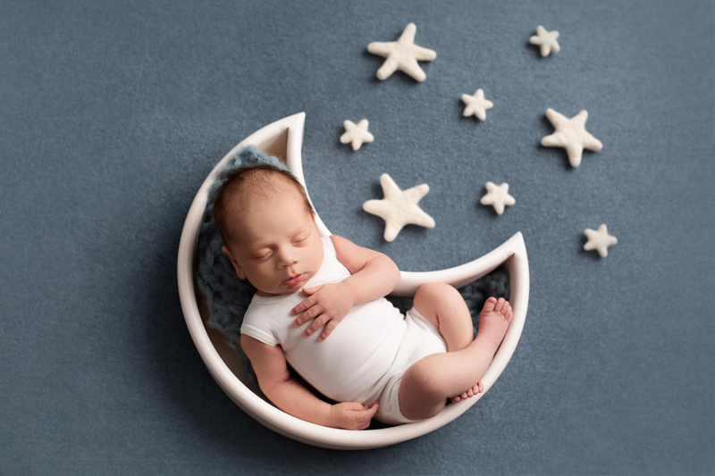 Newborn Photographer, a little baby sleeps inside a container shaped as the crescent moon, seven stars beside him