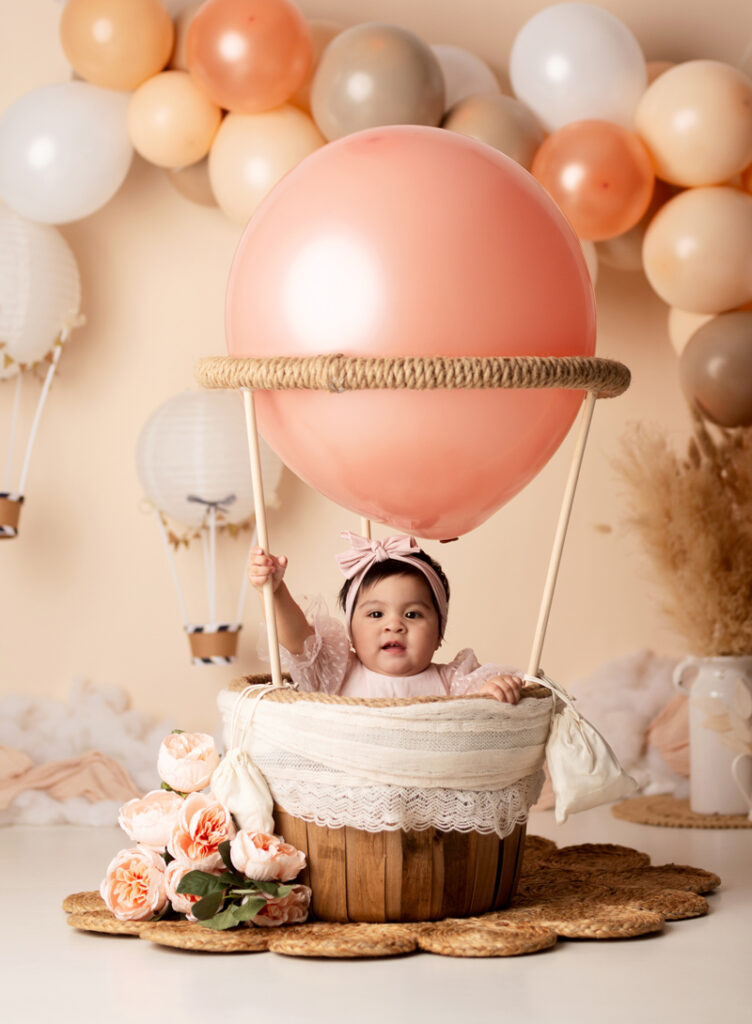 Newborn Photographer, a baby sits in a small basket replica of a hot air balloon, with balloons scattered across the wall behind her