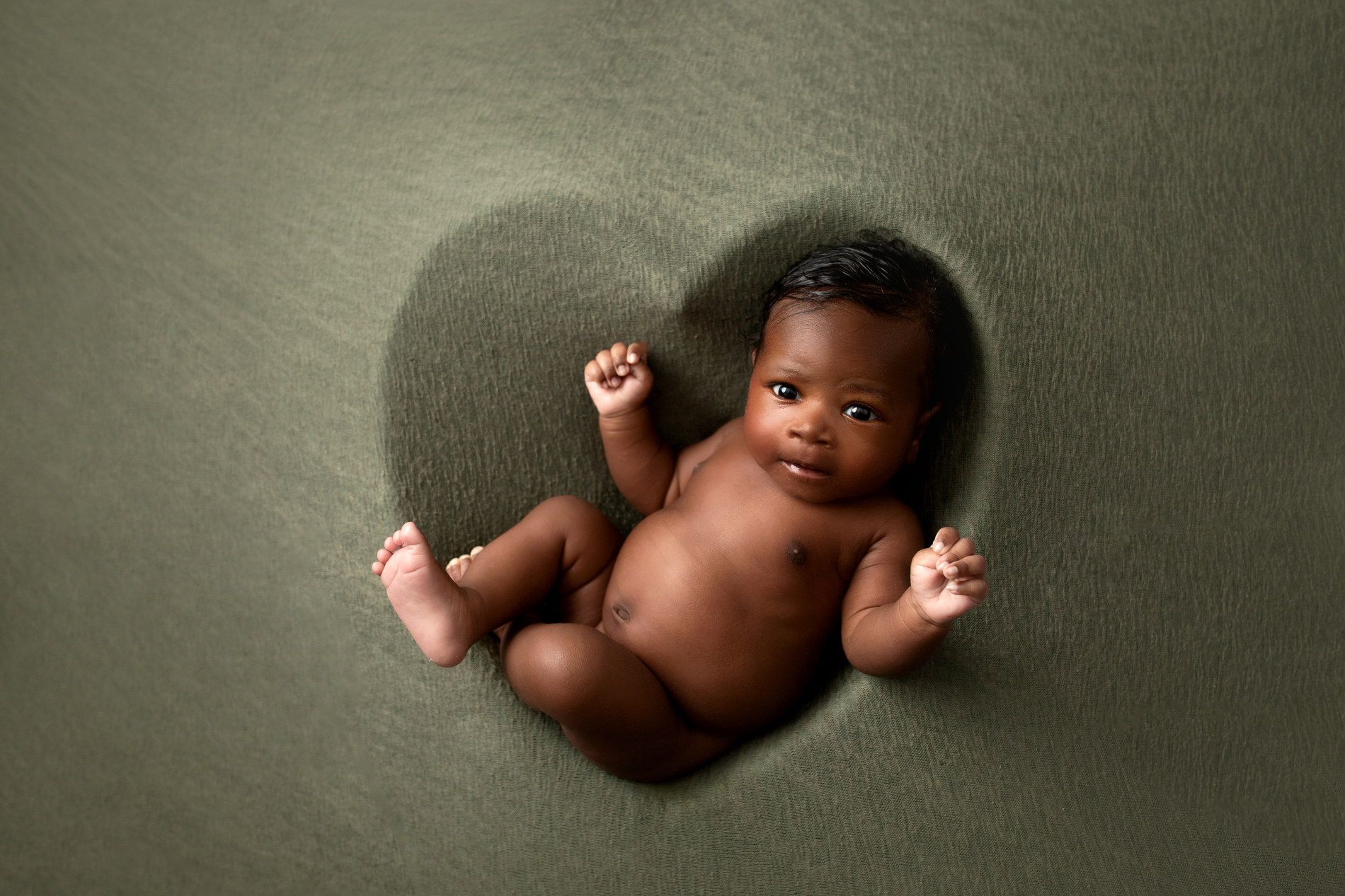 Newborn Photographer, a baby is wide-eyed and cosy on bedsheets, there is an impression of a heart beneath baby