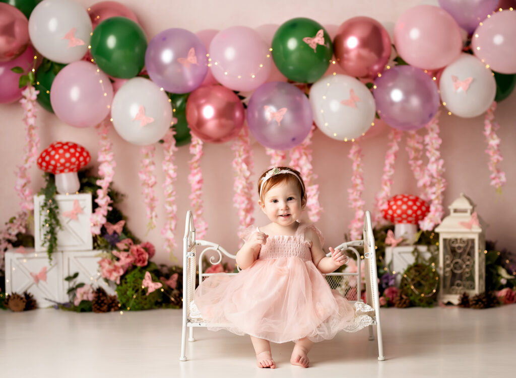 Baby girl smiles on a scroll bench with balloons wearing a pink dress
