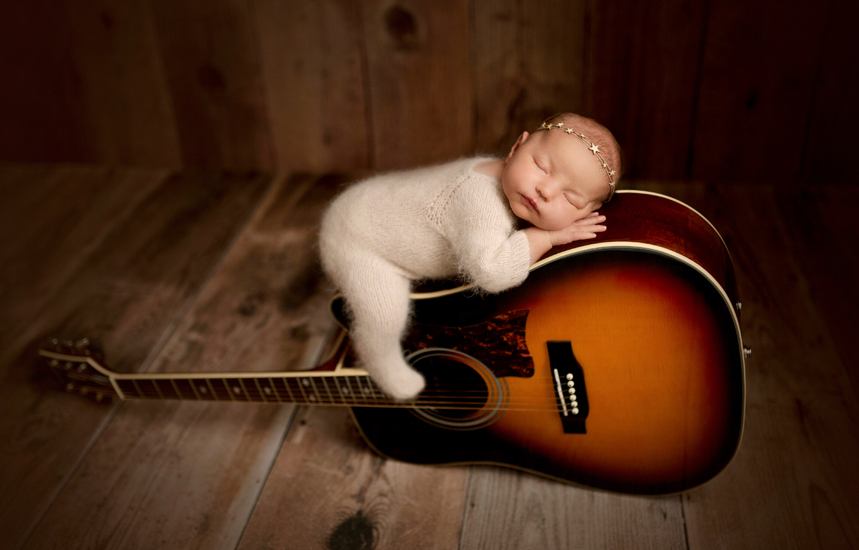 Baby in fuzzy cream outfit posed on guitar on brown wood backdrop 