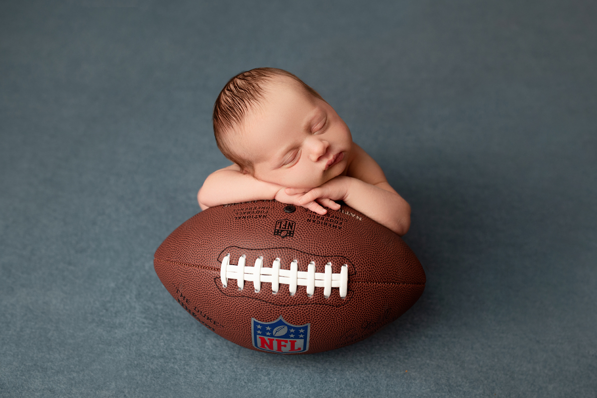 Newborn baby boy posed with his hands under his face on top of a football