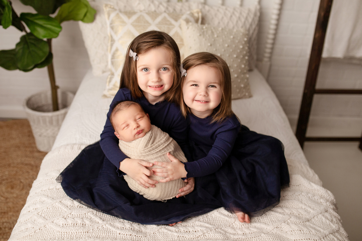 Sisters hold newborn baby brother on a bed wearing navy blue dresses