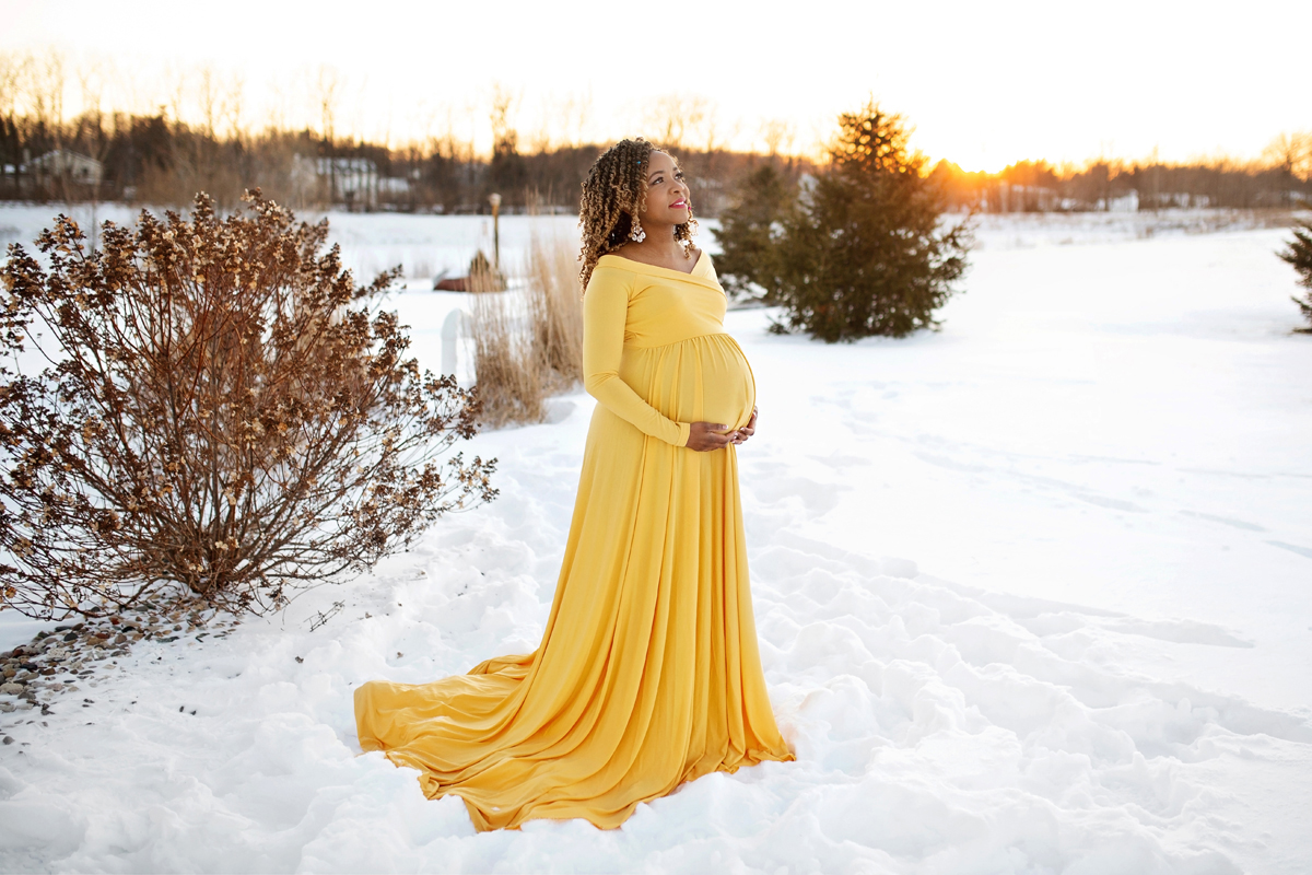 Expecting mother poses in the snow for her maternity photo shoot wearing a yellow dress