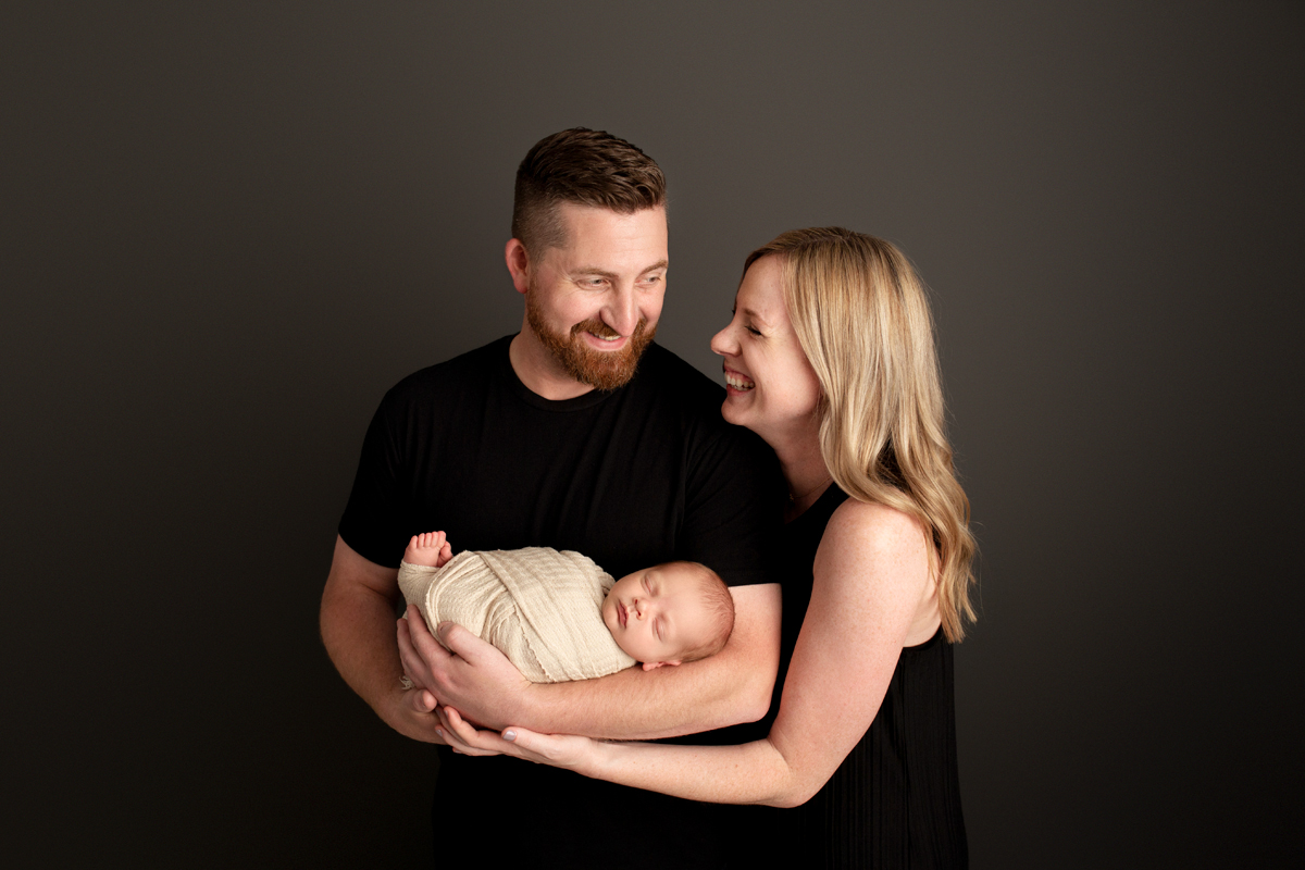 Family smiles with their new baby boy wearing black shirts and baby wears a swaddle wrap