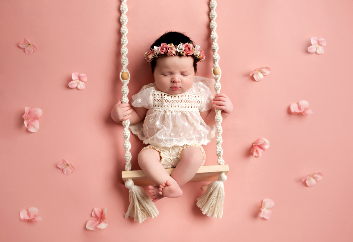 Baby girl sits on a swing with flower crown and flowers around her wearing a cream romper