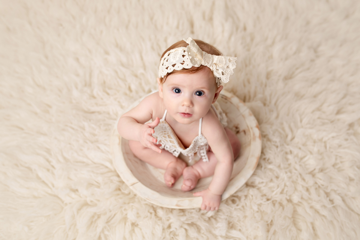 Baby girl sits in a white bowl and looks at the camera wearing a bow in her hair