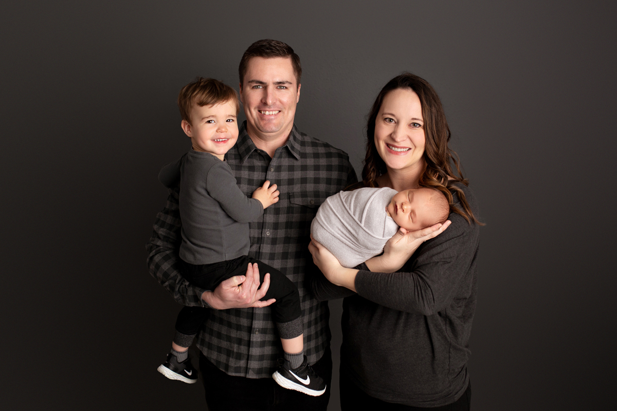 Family poses with newborn and child wearing gray against a gray background