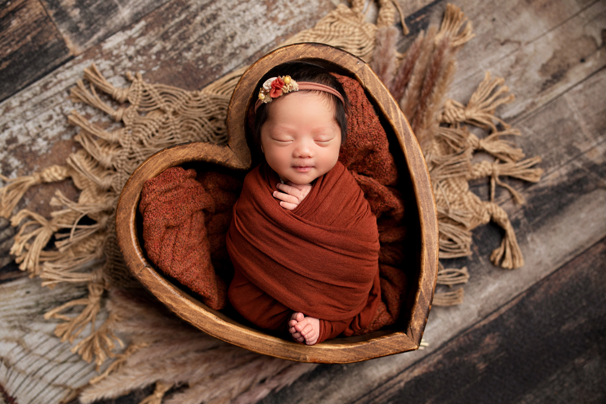 Newborn girl swaddled in rust color wearing a floral headband sleeping in a wooden heart