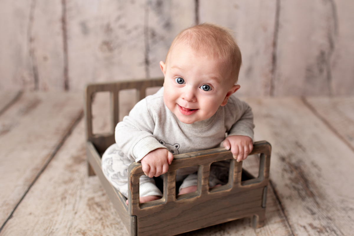Baby Boy Smiles Happily in a Wooden Bed on a Cream Wood Background