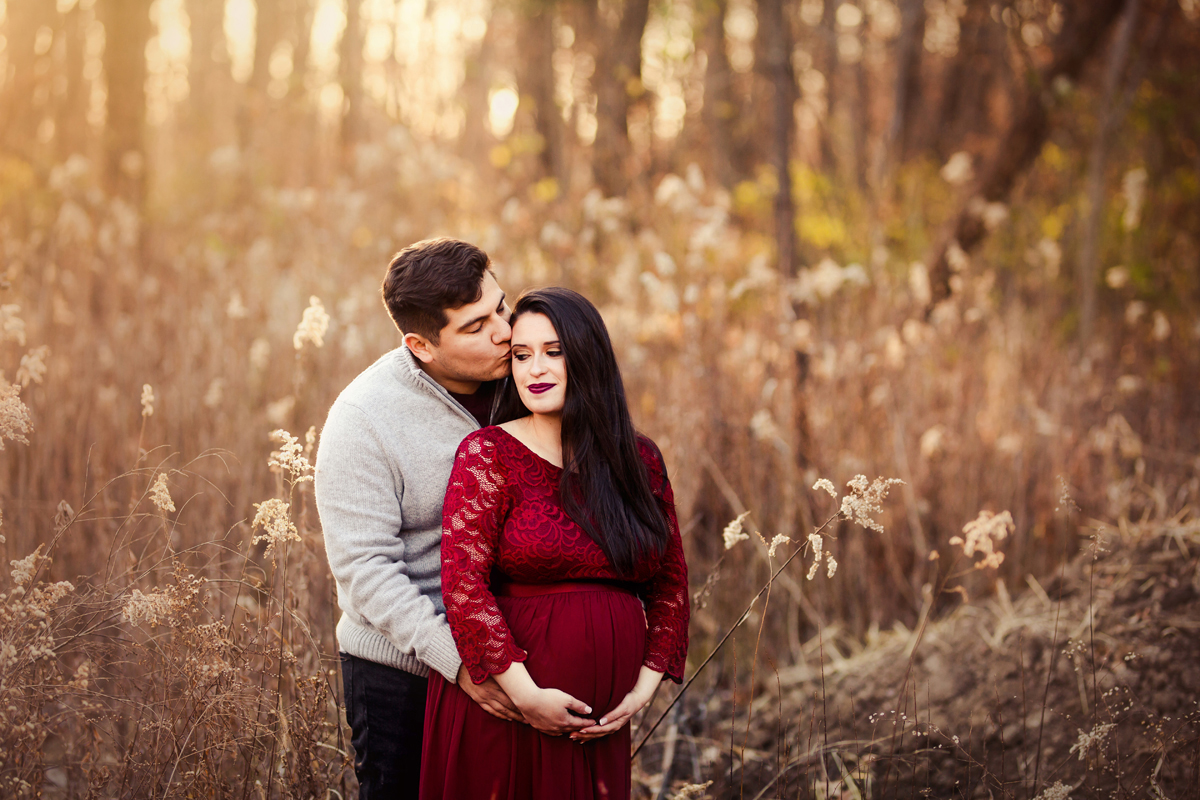 Couple poses in a field with wheatgrass for their maternity session and mother wears a red dress