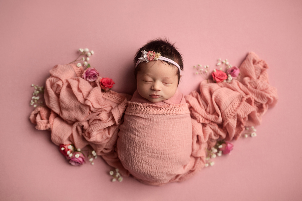 Newborn baby girl sleeps on a blanket with flowers tucked all around her in pink