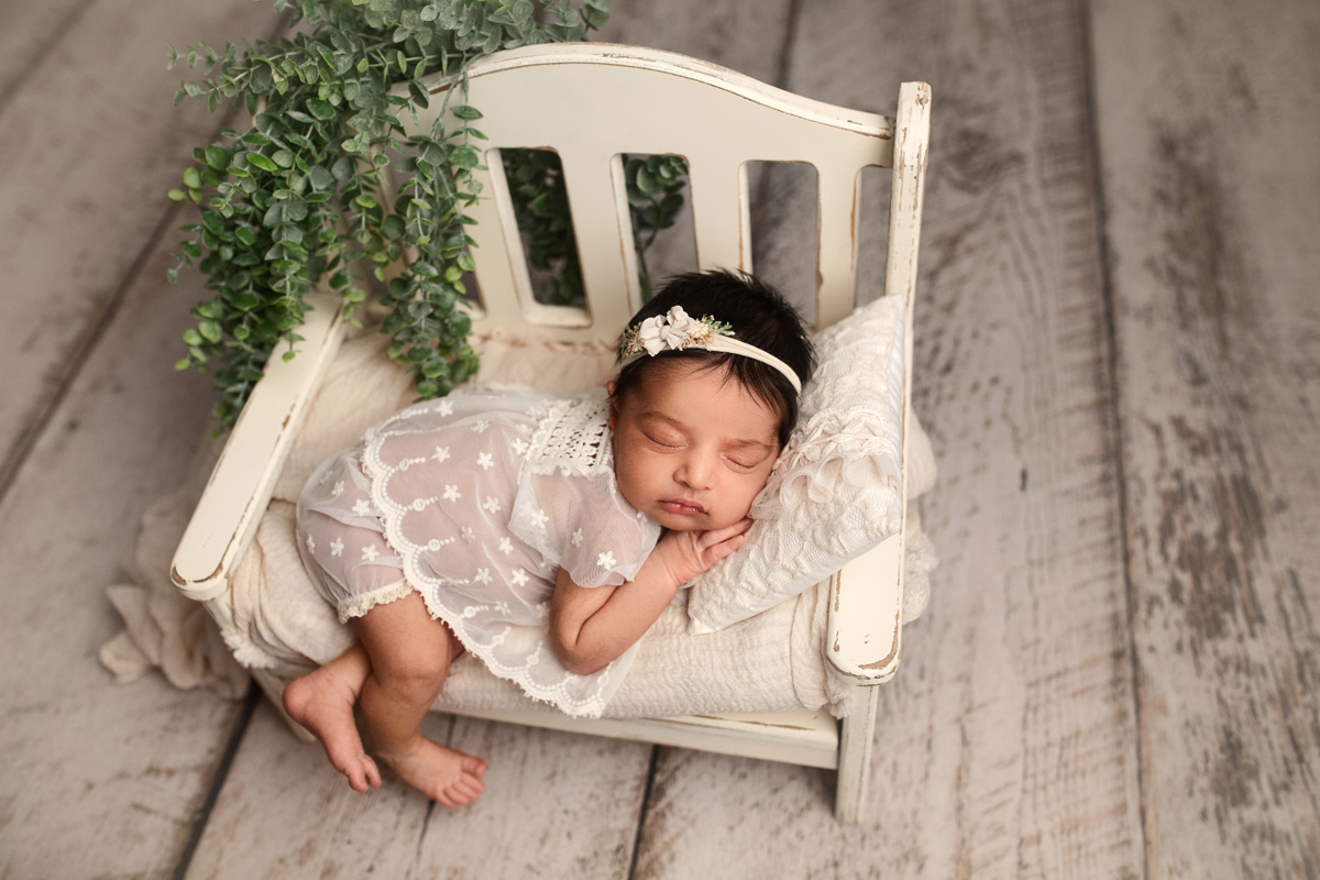 Newborn baby girl wearing a cream lace outfit sleeping on a lace ruffle pillow with greenery