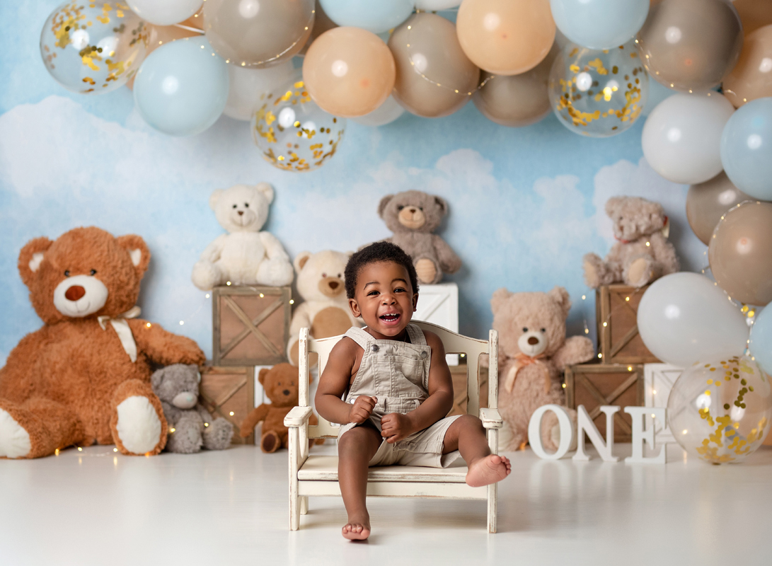 Toddler Boy Sits on White Wooden Bench with Big Smile Surrounded by Big Balloon Banner and Bears