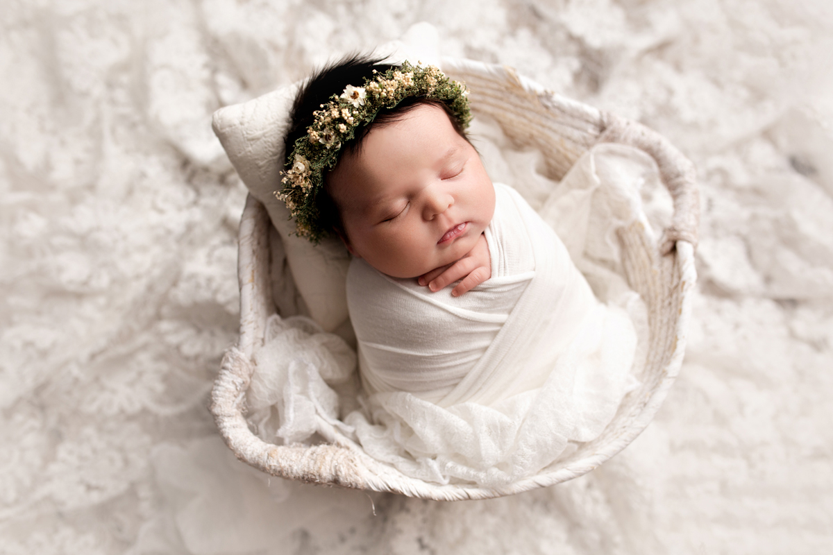 Baby girl is swaddled in a white basket on white lace with a flower crown