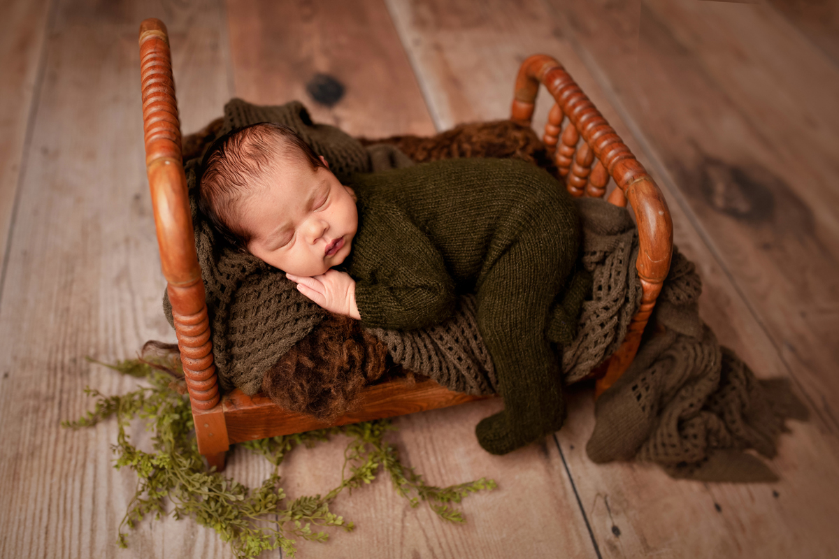 Newborn baby sleeps in a wooden baby bed wearing an olive green knit romper
