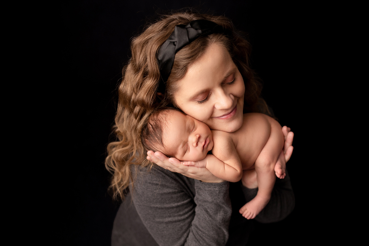 Woman holds new baby girl in her hands with a black background and smiles