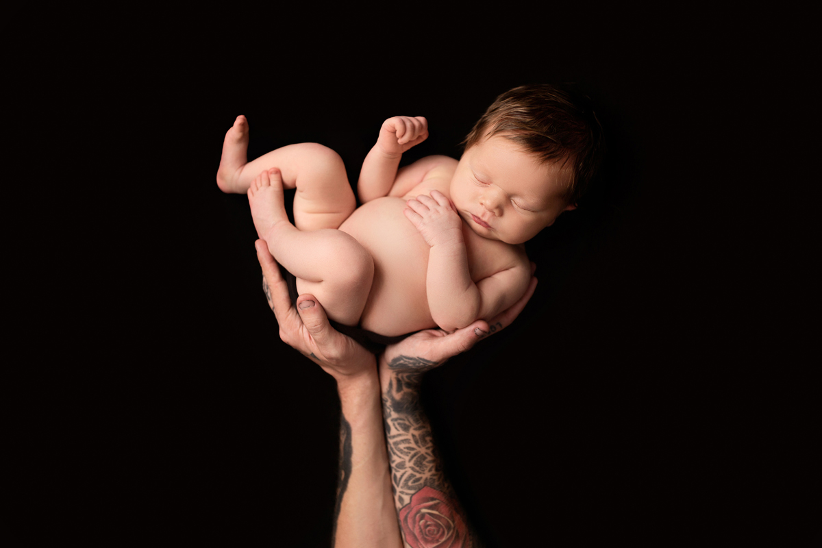 Dad holds new baby girl in the air while she sleeps with a black background and he has a rose tattoo