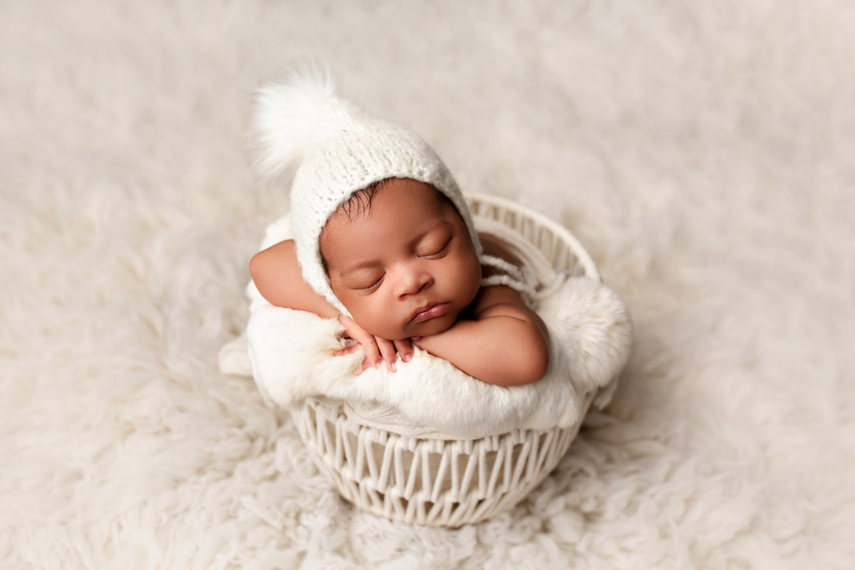 Newborn baby boy is posed in a basket with white fur and a bonnet