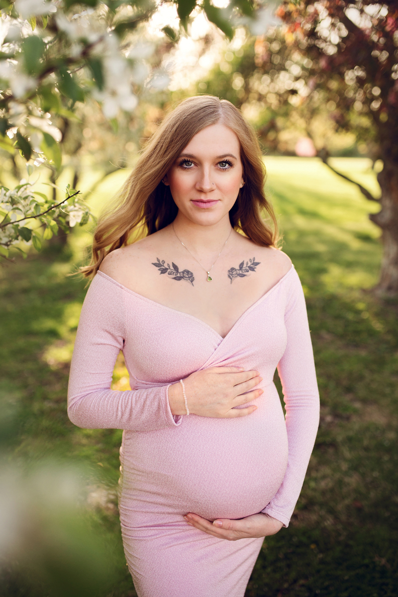 Pregnant woman wears a pink dress and is surrounded by pink flowers and trees