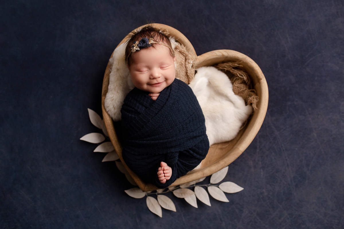 Newborn baby girl smiles in a bowl wearing navy blue swaddle with white leaves
