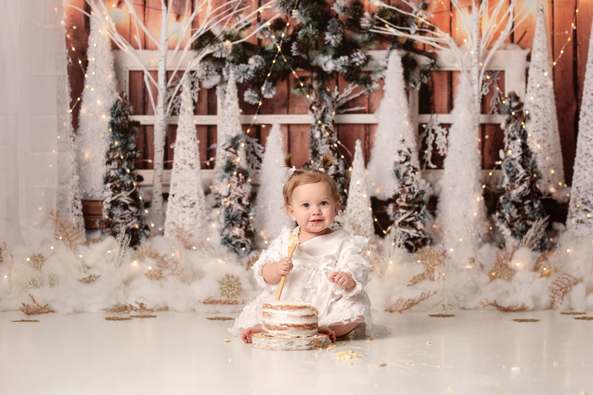 Baby girl holds a spoon and eats cake for her winter wonderland cake smash with Christmas trees