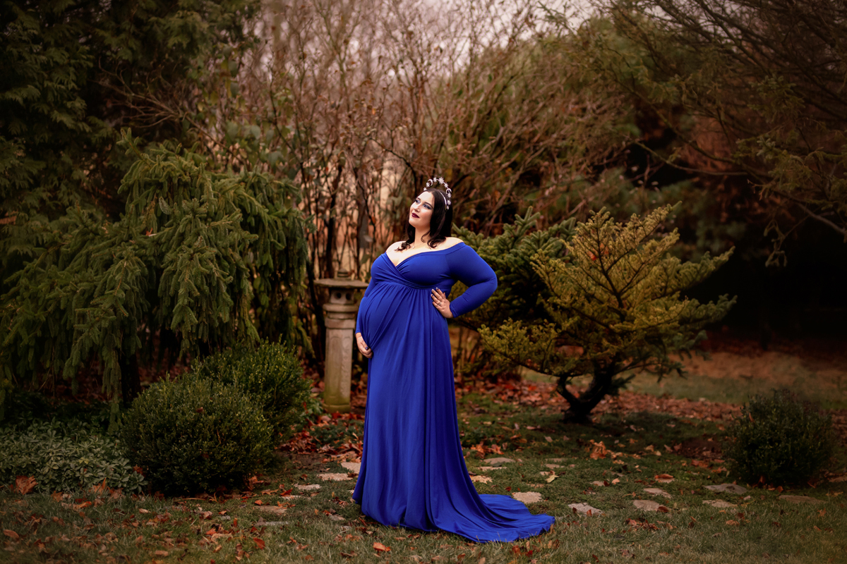 Pregnant woman poses in a garden with a crown on her head wearing a blue maternity dress