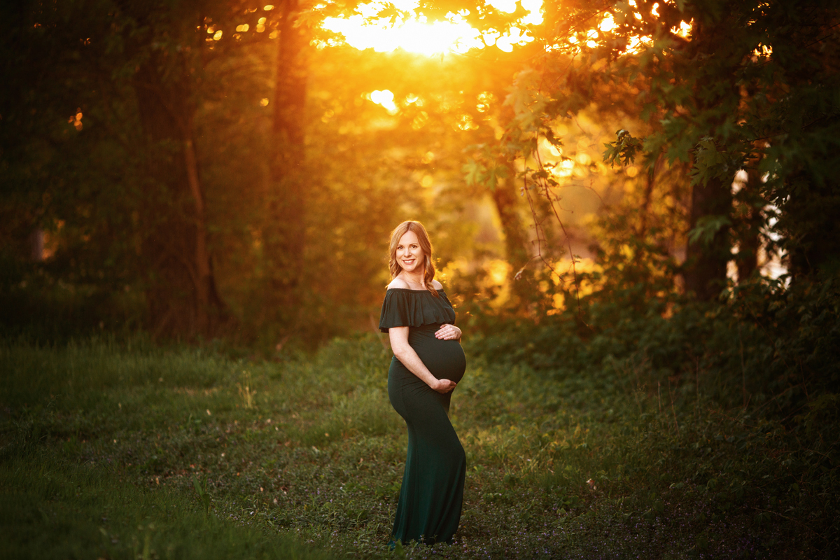 Pregnant woman holds her belly in the grass with the sunset behind her wearing a green maternity gown