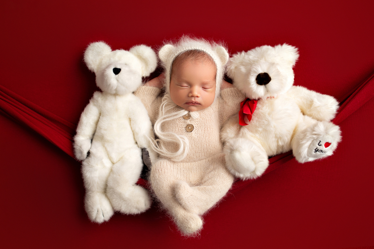 Baby boy sleeps with teddy bears wearing a bear bonnet on red for Valentine's Day