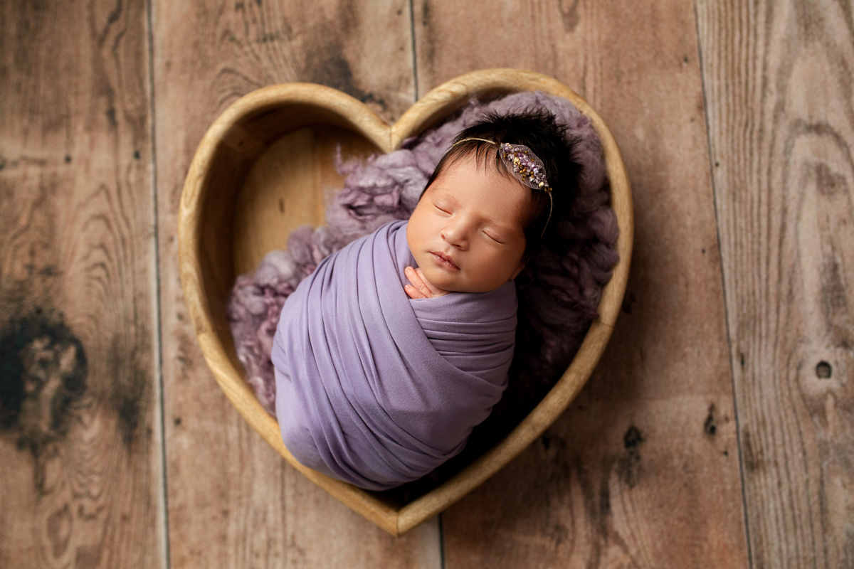 Newborn Girl Swaddled in Purple Wrap in Wooden Heart Bowl on Wood Floor